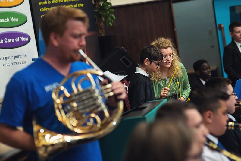 Out of focus in the foreground, a man in a blue Royal Scottish National Orchestra (RSNO) t-shirt plays a french horn while a teacher in the background is helping a pupil play tuned percussion